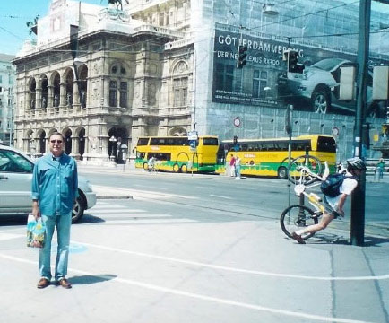 Imagem de um homem a posar para uma fotografia enquanto que atrás dele um ciclista vai contra um poste.